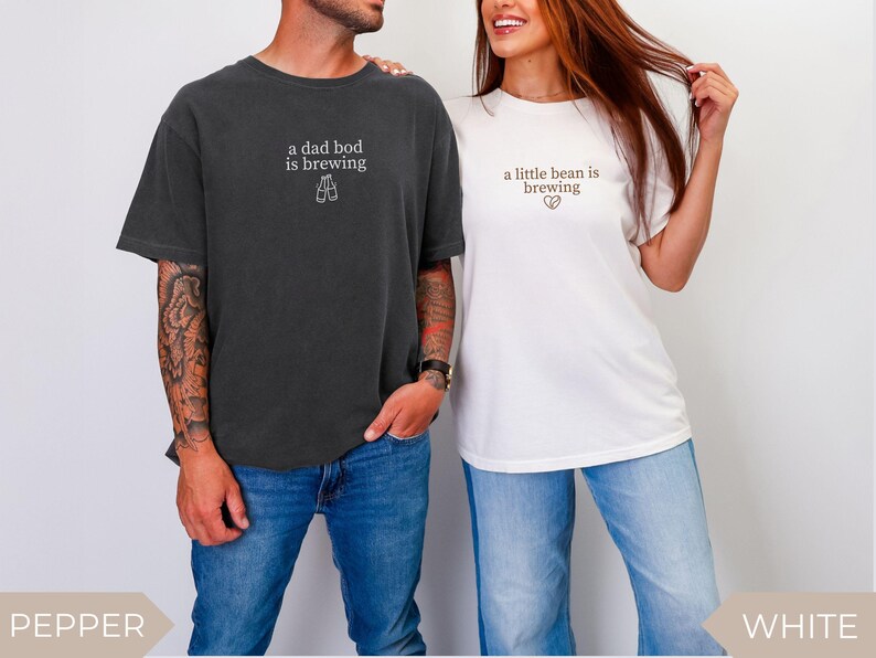 K&ouml;nnte beinhalten: Zwei passende T-Shirts, eines in Dunkelgrau und eines in Wei&szlig;, mit Text. Das dunkelgraue Shirt tr&auml;gt die Aufschrift "a dad bod is brewing" mit einer Bierglas-Grafik. Das wei&szlig;e Shirt tr&auml;gt die Aufschrift "a little bean is brewing" mit einer Herzgrafik. Beide Shirts werden mit Jeans getragen.
