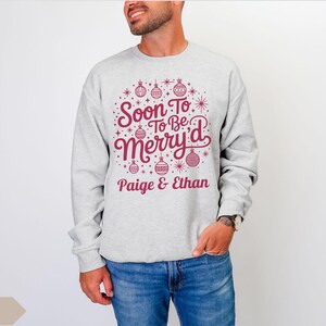 May include: A light gray sweatshirt with the text "Soon To Be Merry'd" in burgundy, along with the names "Paige & Ethan." The design includes Christmas ornaments and stars. The person is wearing blue jeans.