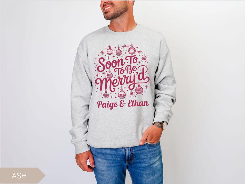 May include: A light gray sweatshirt with the text "Soon To Be Merry'd" in burgundy, along with the names "Paige & Ethan." The design includes Christmas ornaments and stars. The person is wearing blue jeans.