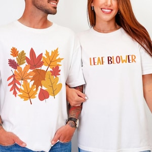 Couples matching fall shirts shown on man and woman. The man’s white tee has a graphic of layered autumn leaves in warm tones of orange, mustard yellow, and burgundy. The woman’s shirt reads “LEAF BLOWER” in bold block letters shaded in fall colors. Both stand side by side, smiling, styled in casual denim jeans. Perfect for autumn outfits, funny couple shirts, fall parties, Thanksgiving gatherings, and cozy seasonal photos.
