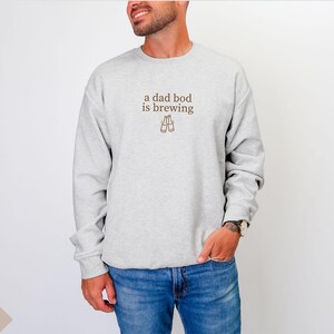 K&ouml;nnte beinhalten: Hellgraues Sweatshirt mit dem braunen Schriftzug "a dad bod is brewing" und zwei Bierflaschen-Illustrationen. Das Model tr&auml;gt eine blaue Jeans. Das Sweatshirt ist ein l&auml;ssiger, bequemer Stil.