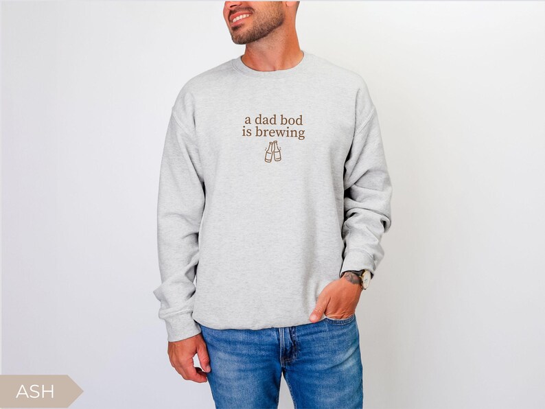 K&ouml;nnte beinhalten: Hellgraues Sweatshirt mit dem braunen Schriftzug "a dad bod is brewing" und zwei Bierflaschen-Illustrationen. Das Model tr&auml;gt eine blaue Jeans. Das Sweatshirt ist ein l&auml;ssiger, bequemer Stil.