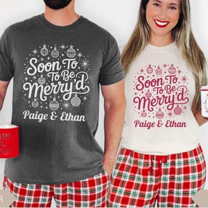 May include: Two people wearing matching Christmas shirts with the text "Soon To Be Merry'd" and the names "Paige & Ethan". The shirts are dark gray and ivory, with red and white text and ornaments. They are holding mugs.