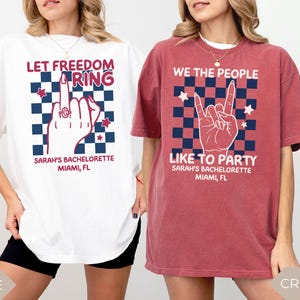 Two women wearing oversized Comfort Colors t shirts in white and crimson colors with patriotic graphic designs for a 4th of July bachelorette party. One shirt reads “Let Freedom Ring” with a hand graphic and custom name and location text. The other shirt reads “We the People Like to Party” with a rock hand gesture design. Styled for a summer girls trip or patriotic bach celebration.
