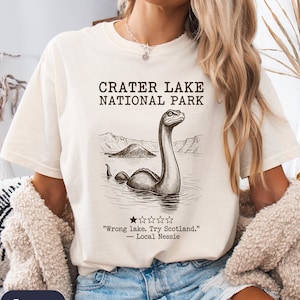 May include: An ivory short-sleeve t-shirt featuring a black graphic of a lake monster in Crater Lake National Park. The text reads: CRATER LAKE NATIONAL PARK, "Wrong lake. Try Scotland." — Local Nessie, with a single star rating icon below.