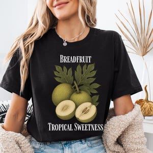 May include: Black t-shirt featuring a breadfruit illustration with green leaves and the text "Breadfruit Tropical Sweetness". The design includes whole and sliced breadfruit. The shirt is a comfortable fit.