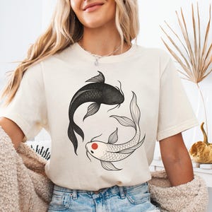 Könnte beinhalten: Cremefarbenes T-Shirt mit einem schwarz-weißen Koi-Fisch-Design. Ein Fisch ist schwarz, der andere weiß mit einem roten Auge, angeordnet im Yin-Yang-Stil. Das Shirt ist ein lässiges Kleidungsstück für den Alltag.