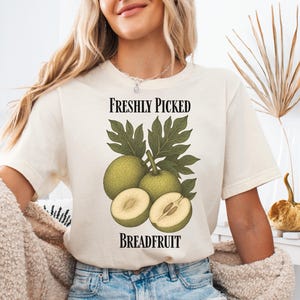 May include: Cream-colored t-shirt featuring a breadfruit illustration with the text "Freshly Picked Breadfruit." The design includes green breadfruit and leaves, with one fruit halved to show the interior. The shirt is a casual, everyday wear item.
