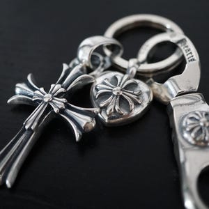 Könnte beinhalten: Ein silberner Schlüsselanhänger mit Kreuz, Herz und Verschluss. Die Kreuz- und Herzanhänger haben aufwendige Blumenmuster. Auf dem Verschluss ist das Wort "hearts" eingraviert. Der Schlüsselanhänger besteht aus glänzendem Metall.