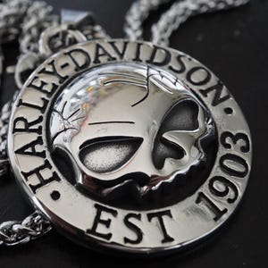 Pode incluir: Colar com pingente Harley-Davidson prateado. O pingente é circular com o logotipo Harley-Davidson e o texto "EST 1903". A corrente é de estilo trançado prateado.