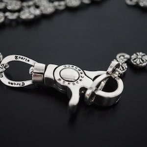 Op de afbeelding: Een zilverkleurige metalen sluiting met het woord "Chrome" gegraveerd, verbonden met een ketting van bloemvormige schakels. De sluiting is het centrale punt, met ingewikkelde details en een gepolijste afwerking. De ketting is gedeeltelijk zichtbaar.