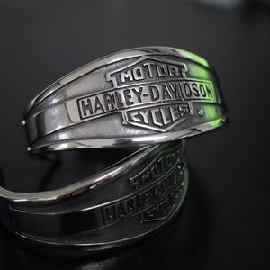 Könnte beinhalten: Zwei silberne Harley-Davidson Armreif-Armbänder. Jedes Armband zeigt das Harley-Davidson-Logo und die Worte "MOTOR HARLEY-DAVIDSON CYCLES" auf der Vorderseite eingraviert. Die Armbänder sind gebogen und haben eine polierte Oberfläche.