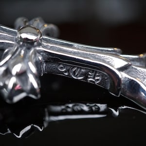 Può includere: Primo piano di un ciondolo a croce color argento con un design floreale dettagliato. Il ciondolo ha una finitura lucida e un piccolo abbellimento rotondo al centro. La croce è appoggiata su una superficie nera riflettente.