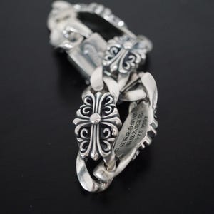 Könnte beinhalten: Ein silbernes Kettenarmband mit verzierten Kreuz- und Blumendesigns. Das Armband hat eine dicke Panzerkette mit dekorativen Gliedern und einem Verschluss. Das Metall ist glänzend und reflektierend, mit graviertem Text auf einem der Glieder.