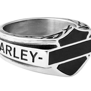 Könnte beinhalten: Ein silberner Ring mit dem Wort "HARLEY-" und einem schwarz-silbernen Emblem. Der Ring hat ein breites Band und ein detailliertes Design auf der Oberseite. Das Emblem hat die Form eines Schildes mit schwarzen Akzenten.