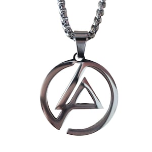 Peut inclure: Un collier pendentif en métal argenté composé d'un cadre circulaire avec une découpe en forme de triangle stylisé au centre. Le pendentif est suspendu à une chaîne épaisse à maillons carrés, présentant une finition métallique polie et brillante.