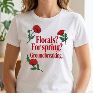 May include: White t-shirt with red floral illustrations and the text "Florals? For spring? Groundbreaking." The design features three red poppy flowers with green leaves. The shirt is a classic crew neck.