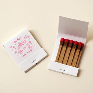 May include: A white matchbook with pink illustrations of party items and the text "Hailey's Last Rodeo". The matchbook is open to reveal a row of brown matches with red tips.
