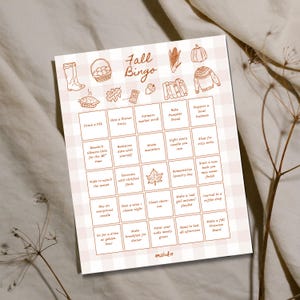 May include: A Fall Bingo game card with a beige and white checked pattern. The card includes fall-themed illustrations and activities such as "Drink a PSL" and "Make a fall Pinterest board". The words "Fall Bingo" are at the top.