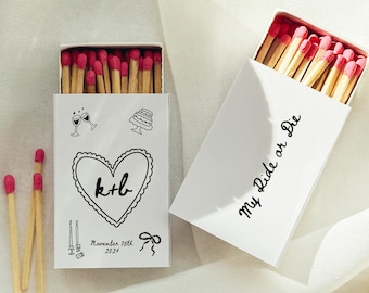 May include: White matchboxes with red-tipped matches. One box features a heart with "k+b" and the date "November 15th 2024". The other box reads "My Ride or Die". Wedding favors.