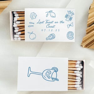 以下が含まれることがあります： 青いイラストとテキストが入った白いマッチ箱が2つ。1つの箱には、海岸をテーマにしたイラストと「Last Toast on the Coast 07.12.25」の文字が入っています。もう1つの箱にはカクテルグラスのイラストが入っています。