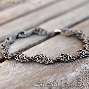 May include: A silver chain bracelet with a woven design. The bracelet is lying on a wooden surface.