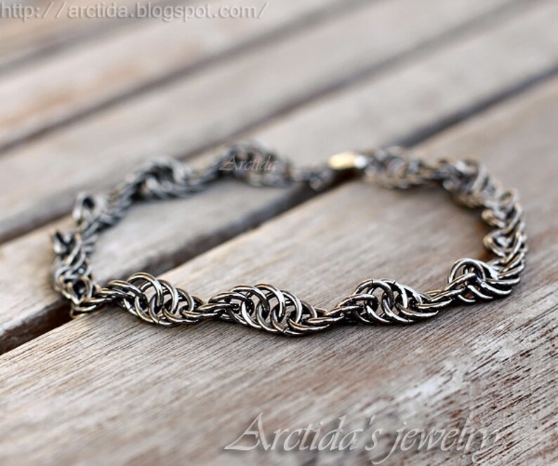 May include: A silver chain bracelet with a woven design. The bracelet is lying on a wooden surface.