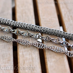 May include: Three silver metal chains of different styles and sizes lay on a wooden surface. The chains are made of small, interlocking links.