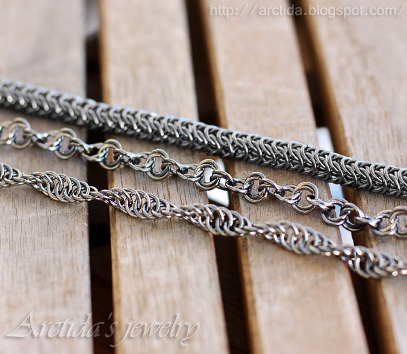 May include: Three silver metal chains of different styles and sizes lay on a wooden surface. The chains are made of small, interlocking links.