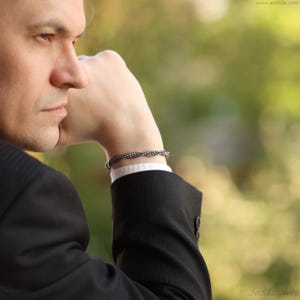 May include: A man wearing a black suit jacket with a white shirt underneath. He is wearing a silver bracelet on his left wrist.