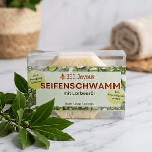 Könnte beinhalten: Ein Badeschwamm in einer transparenten Kunststoffbox. Auf dem Etikett steht "BEE Joyous SEIFENSCHWAMM mit Lorbeeröl Bath Soap Sponge". Der Schwamm ist hellbeige und hat eine geometrische Form. Das Etikett weist auch "2 in 1 Reinigung & Peeling" aus.