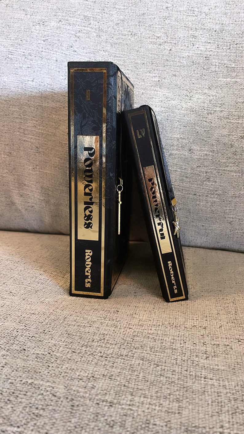 May include: Two black hardcover books with gold lettering and gold bookmarks. The books are titled "Powerless" and "Powerful" by Roberts.