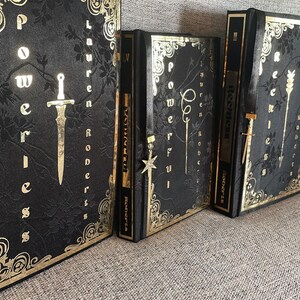 May include: Three black hardback books with gold accents and titles. The books are titled "Powerless", "Prophecy", and "Reckless".