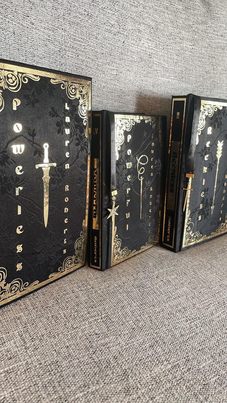 May include: Three black hardback books with gold accents and titles. The books are titled "Powerless", "Prophecy", and "Reckless".