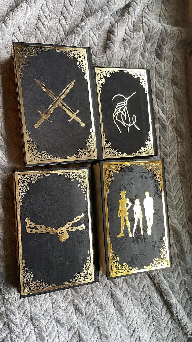 Op de afbeelding: Vier zwarte boeken met gouden randen en decoratieve ontwerpen. De omslagen tonen gekruiste zwaarden, een zweep, een ketting met een hangslot en drie figuren.