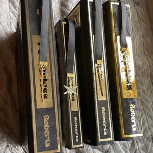 Op de afbeelding: Vier zwarte hardcover boeken met gouden accenten en gouden lintjes als bladwijzers. Elk bladwijzer heeft een andere gouden hanger. De boeken zijn getiteld "Roberts".