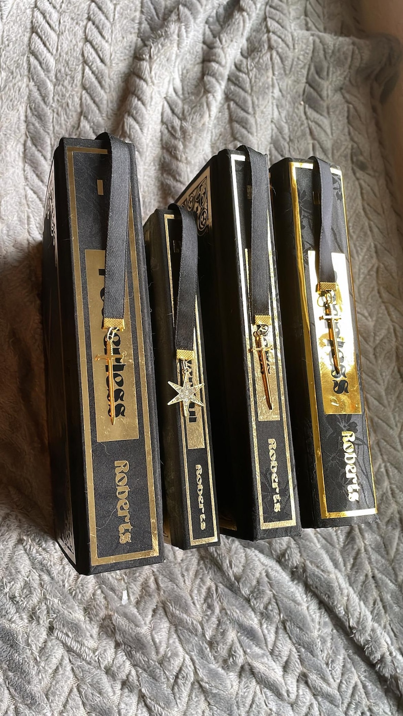 Op de afbeelding: Vier zwarte hardcover boeken met gouden accenten en gouden lintjes als bladwijzers. Elk bladwijzer heeft een andere gouden hanger. De boeken zijn getiteld "Roberts".