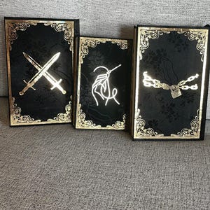 May include: Three black journals with gold trim and decorative designs. The first journal has a gold sword design, the second has a gold needle and thread design, and the third has a gold chain and padlock design.