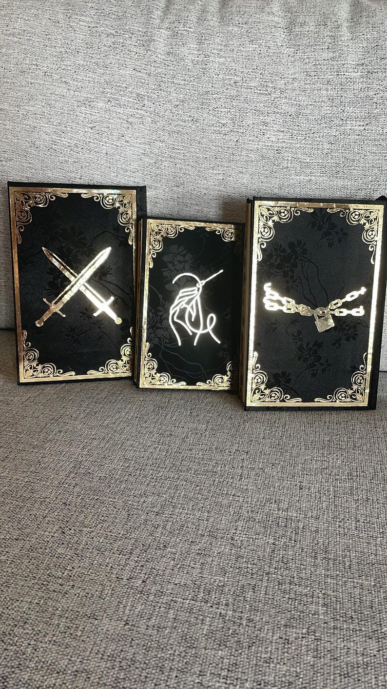 May include: Three black journals with gold trim and decorative designs. The first journal has a gold sword design, the second has a gold needle and thread design, and the third has a gold chain and padlock design.