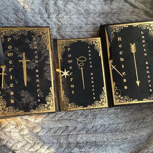 May include: Three black hardback journals with gold accents and decorative bookmarks. The journals are titled "Powerful", "Reckless", and "Brave".