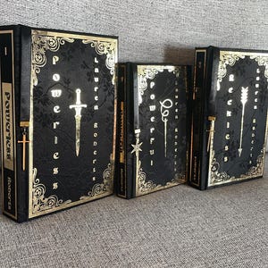 May include: Three black hardback books with gold accents and decorative designs. The books are titled "Powerless", "Powerful", and "Reckless" and are by Lauren Roberts.