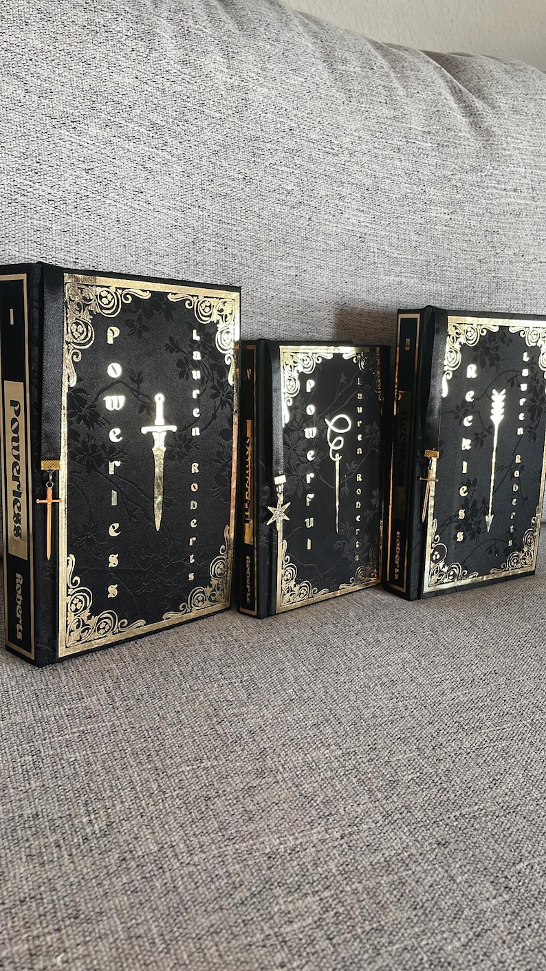 May include: Three black hardback books with gold accents and decorative designs. The books are titled "Powerless", "Powerful", and "Reckless" and are by Lauren Roberts.