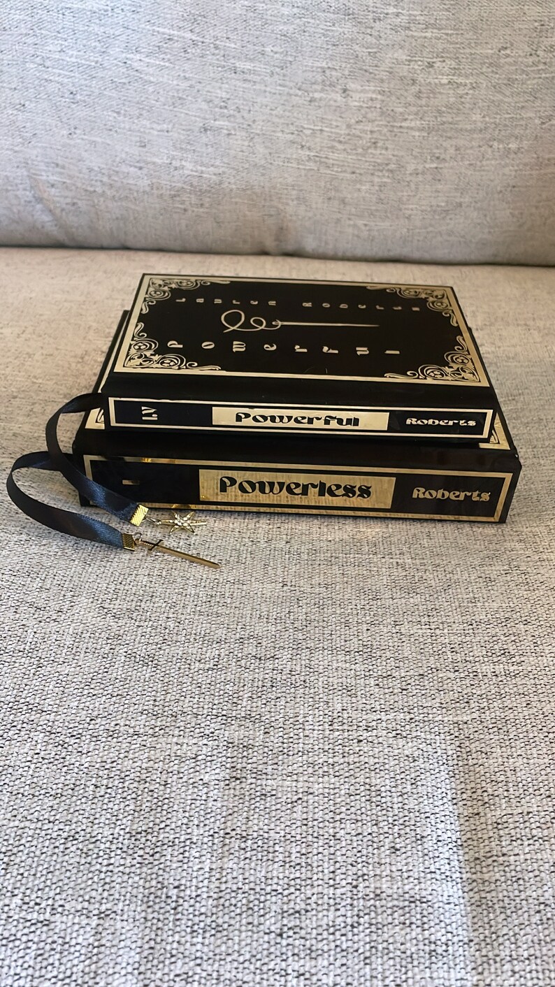 May include: Two black hardcover books with gold lettering. The top book is titled "Powerful" by Roberts. The bottom book is titled "Powerless" by Roberts. Both books have black ribbon bookmarks.