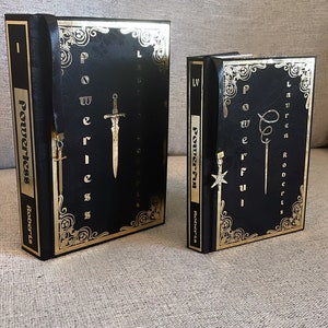 May include: Two black hardcover books with gold accents and the word "Powerless" and "Powerful" on the spines. The books have gold bookmarks with a sword and a star design.