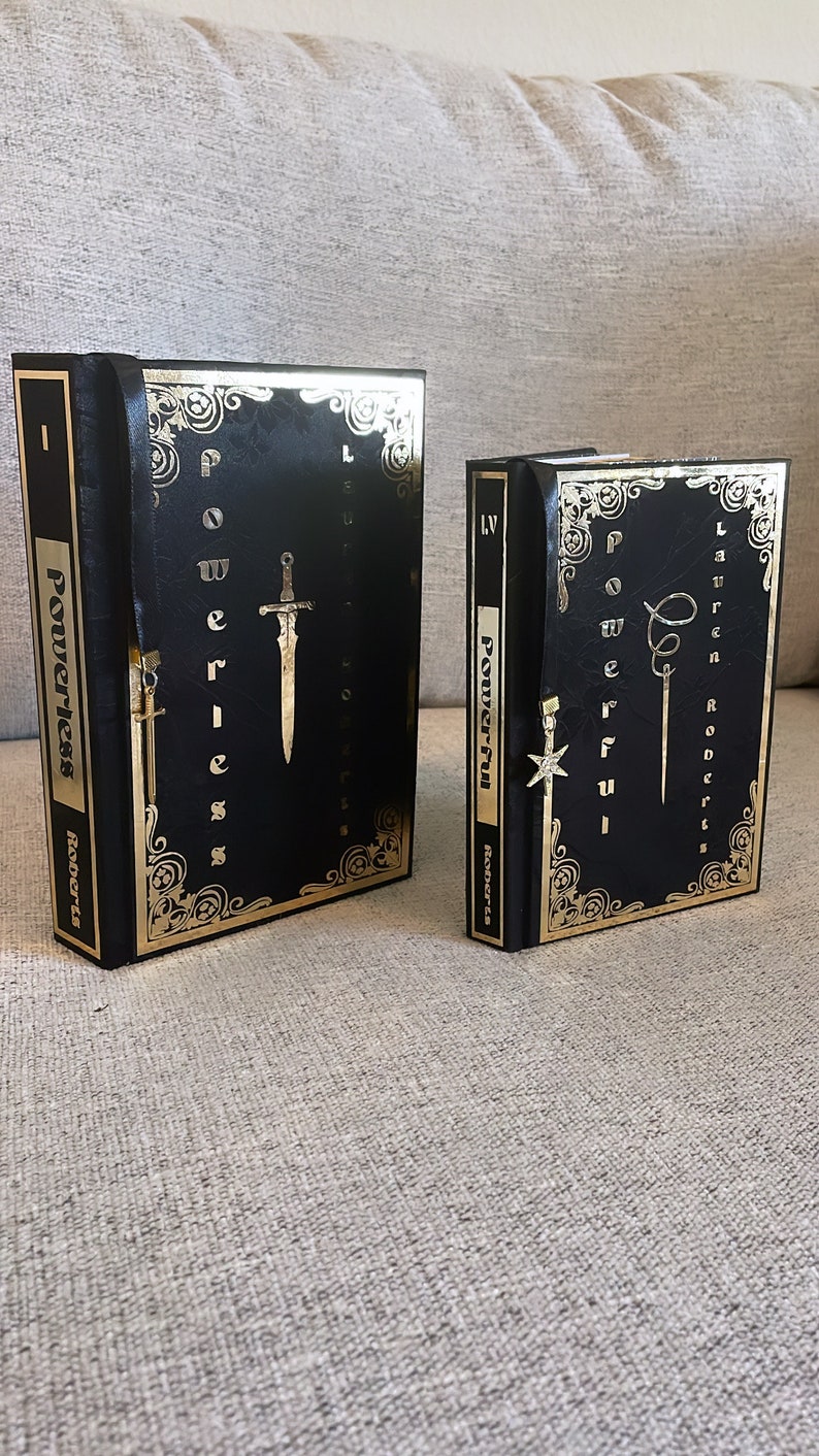 May include: Two black hardcover books with gold accents and the word "Powerless" and "Powerful" on the spines. The books have gold bookmarks with a sword and a star design.