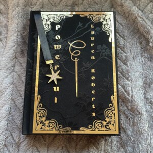 May include: A black book with gold accents and a black bookmark with a gold star charm. The book's title is "Powerful" and the author is "Lauren Roberts".