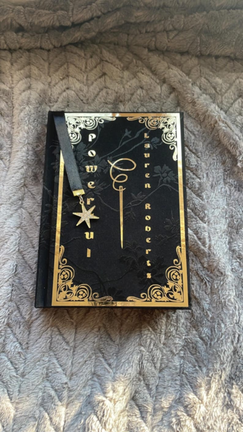May include: A black book with gold accents and a black bookmark with a gold star charm. The book's title is "Powerful" and the author is "Lauren Roberts".