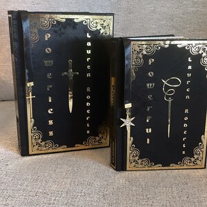 May include: Two black hardcover books with gold accents and decorative bookmarks. The books are titled "Power of Weakness" and "Power of Fierceness" by Lauren Roberts.