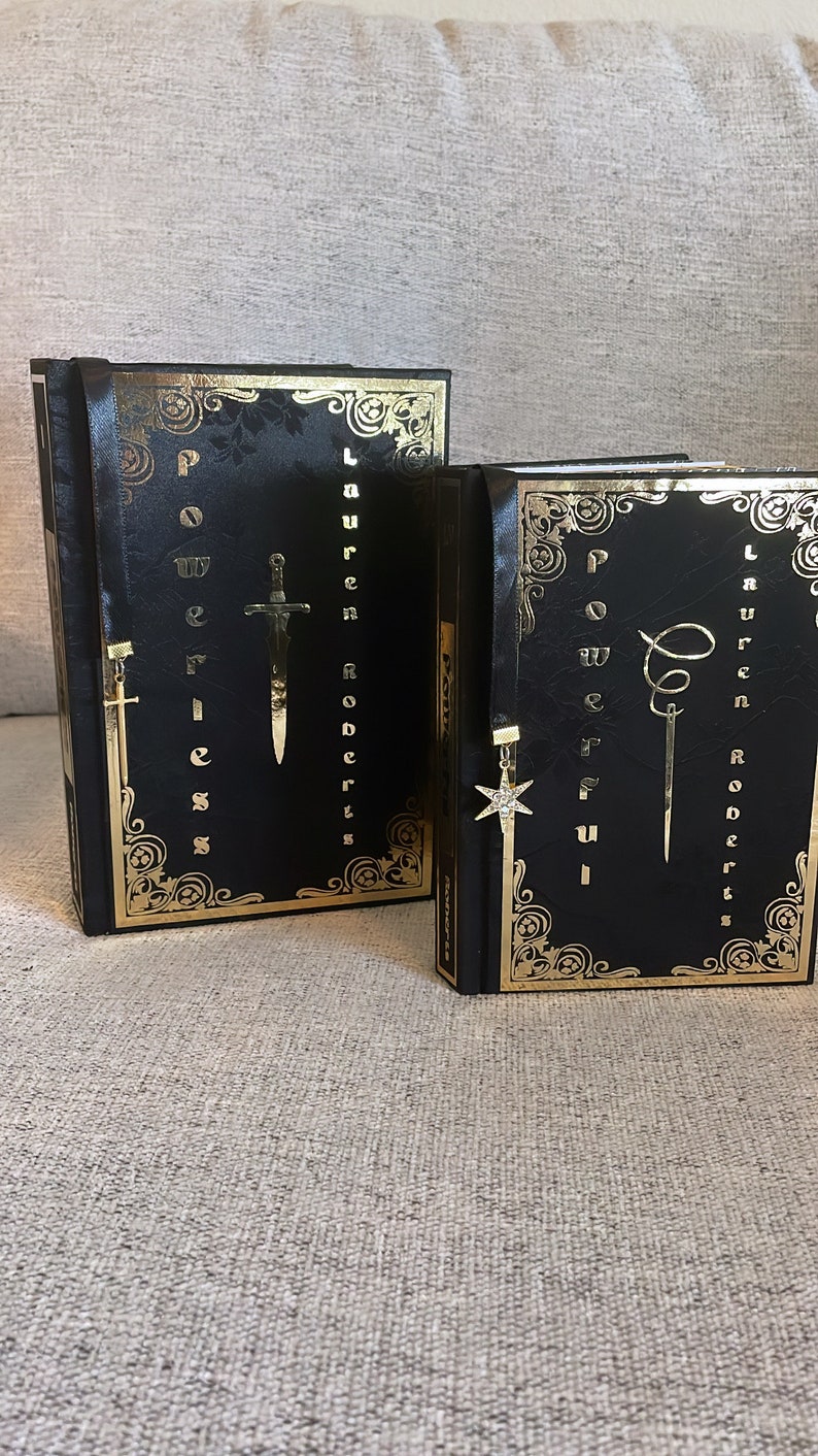 May include: Two black hardcover books with gold accents and decorative bookmarks. The books are titled "Power of Weakness" and "Power of Fierceness" by Lauren Roberts.