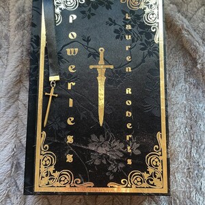 May include: A black book with a gold-coloured design and the title "Powerless" by Lauren Roberts. A gold-coloured sword and cross are on the cover.